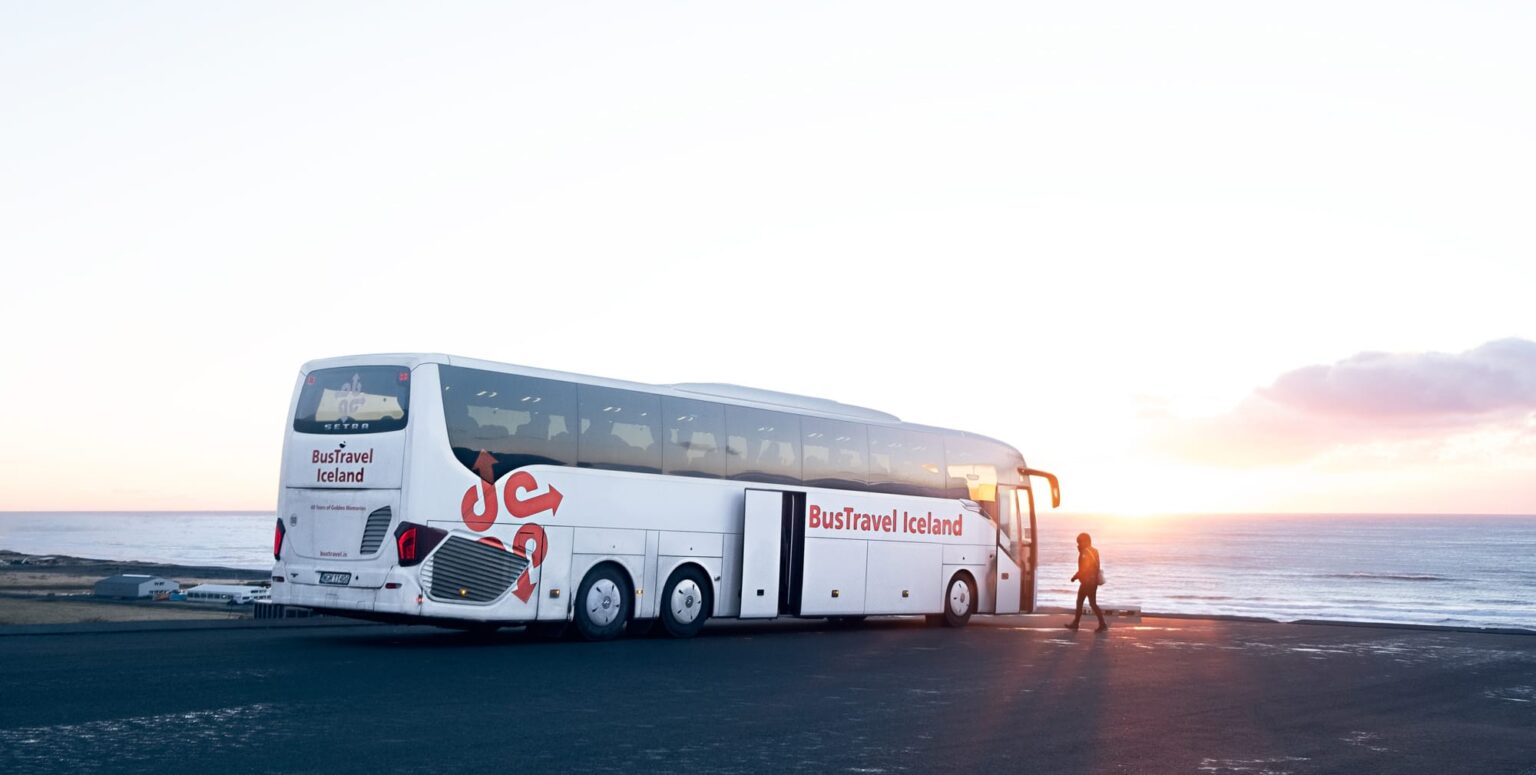 Biały autokar firmy BusTravel Iceland zaparkowany na surowej, ciemnej drodze wybrzeża w Islandii o zachodzie słońca. Jedna osoba idzie w kierunku otwartych drzwi autobusu.